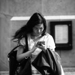 Black and white photo of young woman looking down at a cell phone; she wears a shoulder bag and carries a coat
