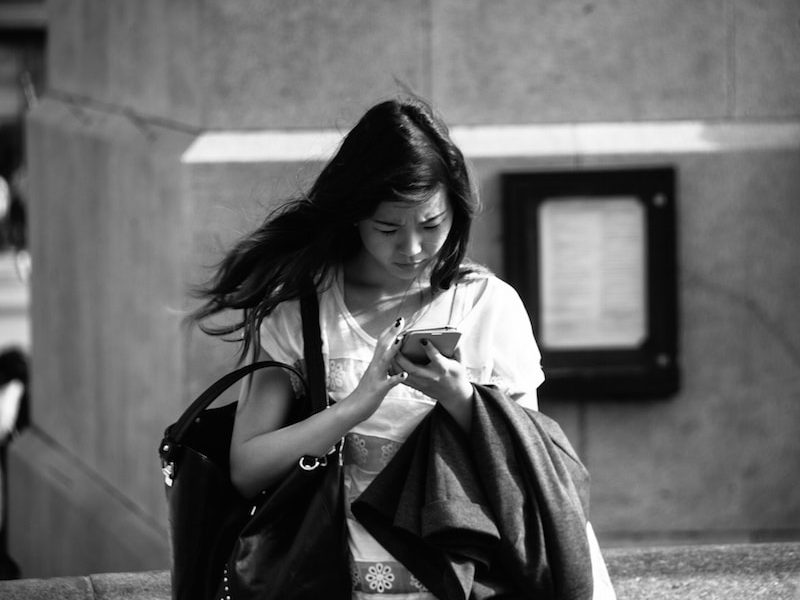 Black and white photo of young woman looking down at a cell phone; she wears a shoulder bag and carries a coat