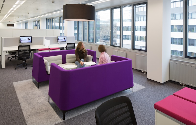 Photo showing young women seated on purple couches facing each other in windowed office building