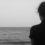 Black and white photo of a woman, shown from back, looking out to sea; pensive mood