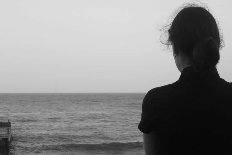 Black and white photo of a woman, shown from back, looking out to sea; pensive mood